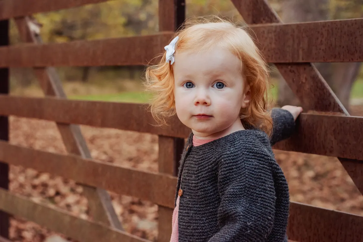 Red-haired toddler beside a rustic fence in fall