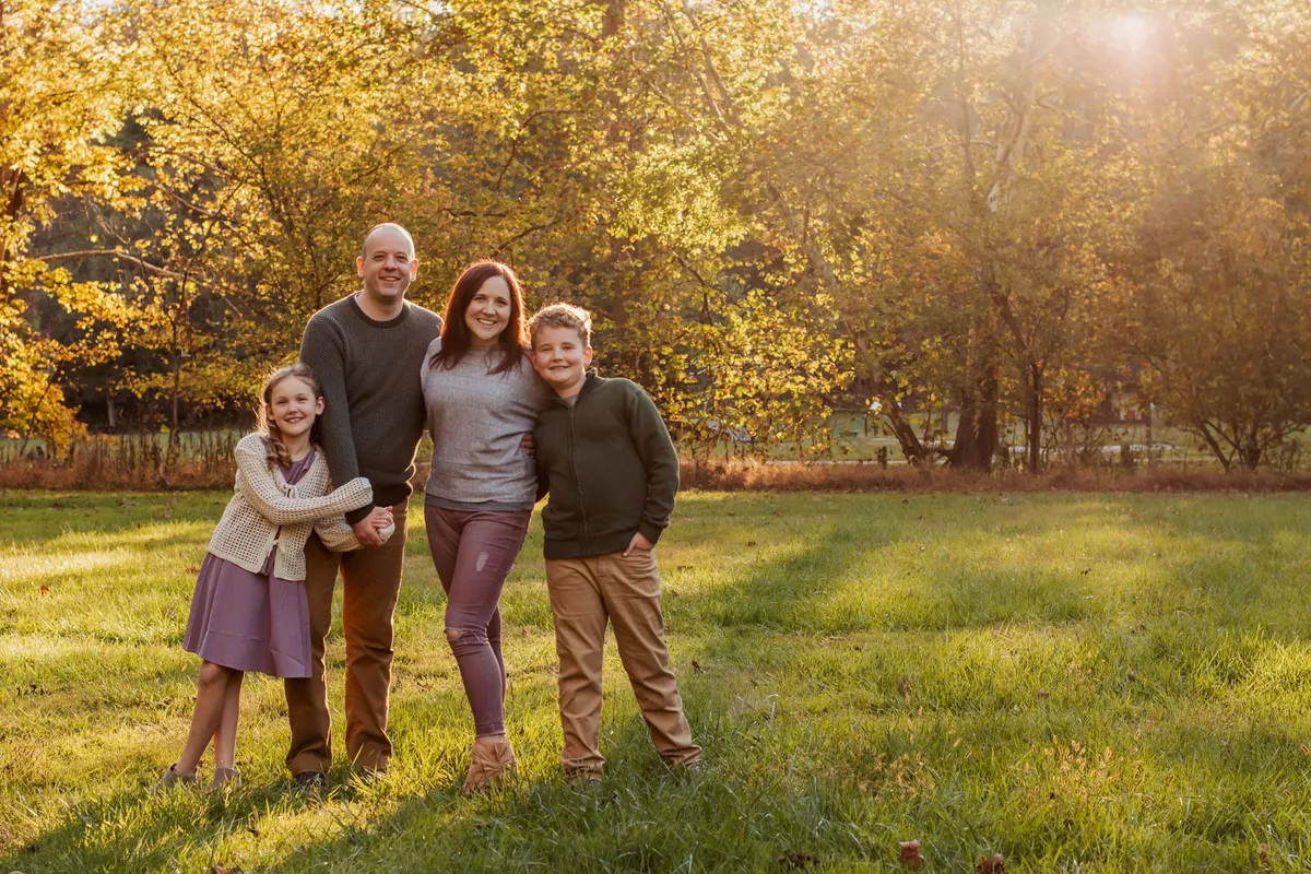 Family of four standing together in golden-hour light with fall trees behind them